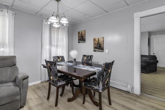 dining space featuring light wood finished floors, a drop ceiling, a chandelier, a baseboard heating unit, and baseboards