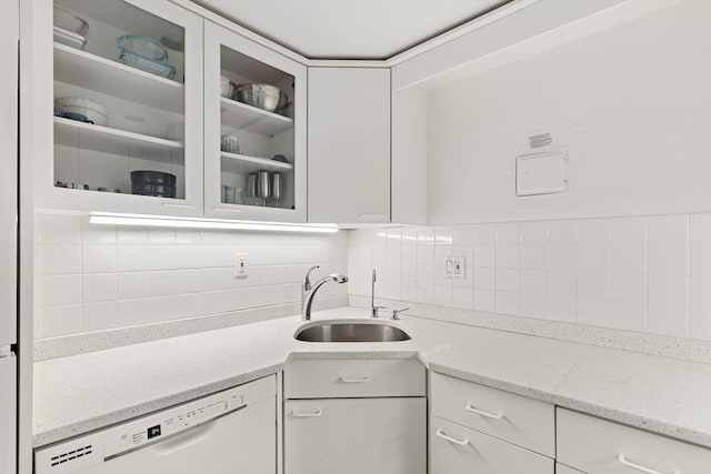 kitchen with white dishwasher, a sink, white cabinets, glass insert cabinets, and tasteful backsplash