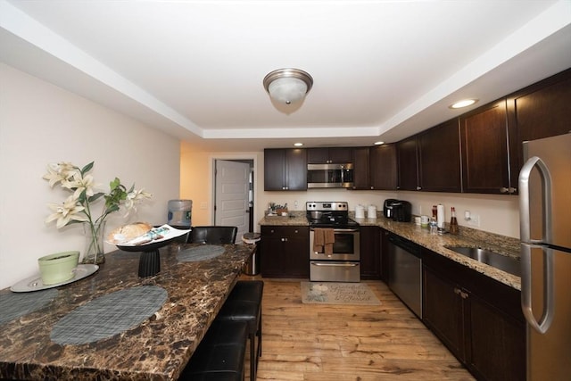 kitchen featuring dark stone counters, light wood-style flooring, stainless steel appliances, dark brown cabinetry, and a raised ceiling