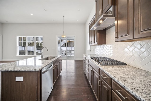 kitchen with dark brown cabinetry, decorative light fixtures, an island with sink, stainless steel appliances, and light stone countertops