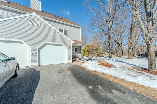 view of snow covered exterior featuring driveway, a shingled roof, a chimney, and an attached garage