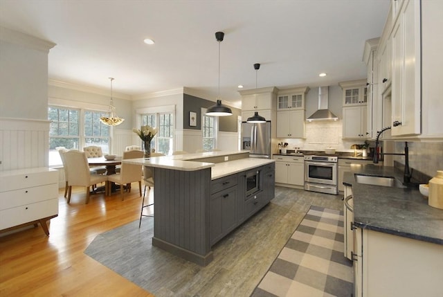 kitchen with a wainscoted wall, stainless steel appliances, wall chimney range hood, and a sink