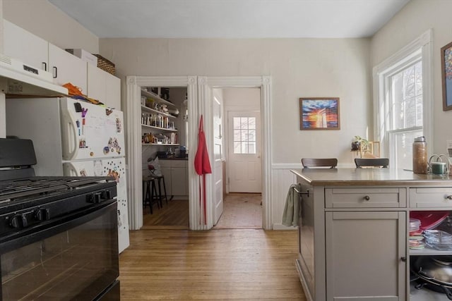 kitchen featuring black gas stove, wainscoting, freestanding refrigerator, under cabinet range hood, and light wood-style flooring