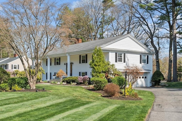 raised ranch featuring asphalt driveway, an attached garage, a chimney, a front lawn, and covered porch
