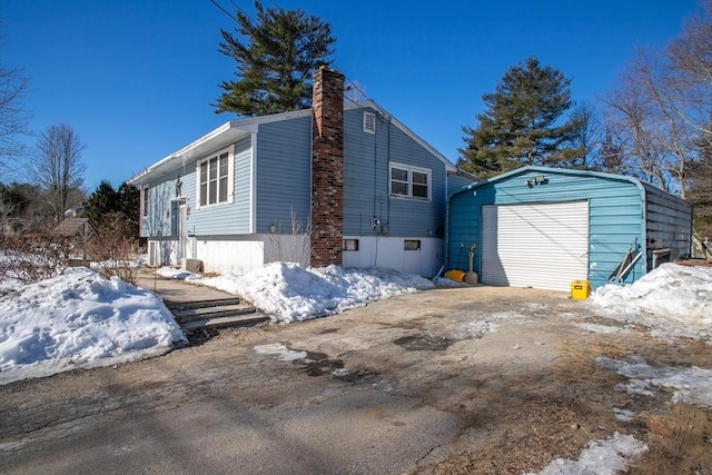 snow covered property with a garage, an outbuilding, a chimney, and driveway