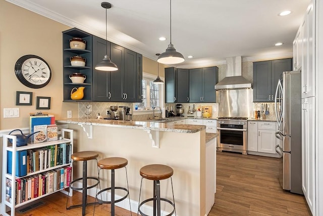 kitchen featuring light stone countertops, a peninsula, stainless steel appliances, wall chimney exhaust hood, and dark wood-style flooring