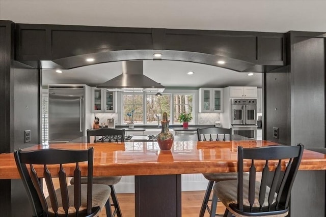 279 Norfolk St, Holliston, MA 01746 - kitchen with appliances with stainless steel finishes, light wood-style flooring, glass insert cabinets, white cabinetry, and wall chimney range hood