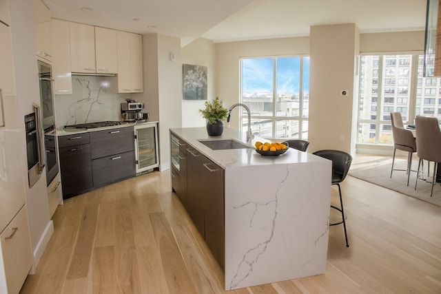 kitchen with dark brown cabinetry, beverage cooler, sink, white cabinets, and an island with sink