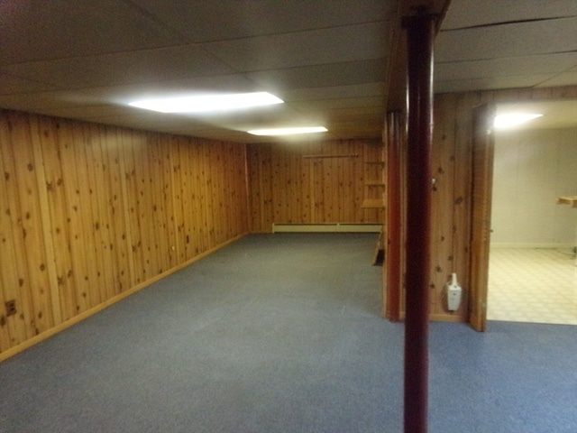 basement featuring a paneled ceiling, wooden walls, baseboard heating, and carpet floors