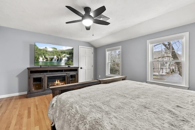 bedroom with baseboards, ceiling fan, vaulted ceiling, light wood-style floors, and a glass covered fireplace