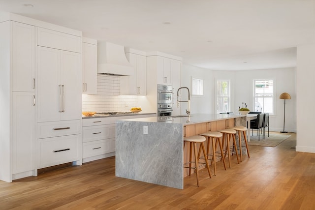 kitchen with white cabinets, premium range hood, a kitchen island with sink, and light hardwood / wood-style flooring