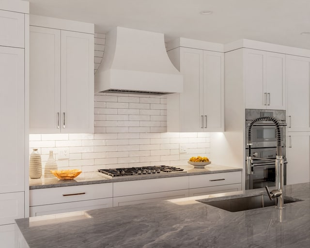 kitchen featuring decorative backsplash, white cabinets, stone countertops, stainless steel appliances, and premium range hood