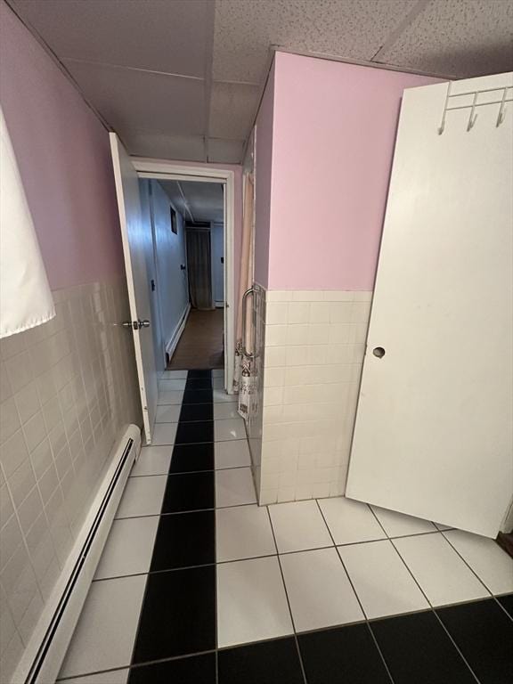 hallway with a wainscoted wall, tile walls, a baseboard heating unit, light tile patterned flooring, and a drop ceiling