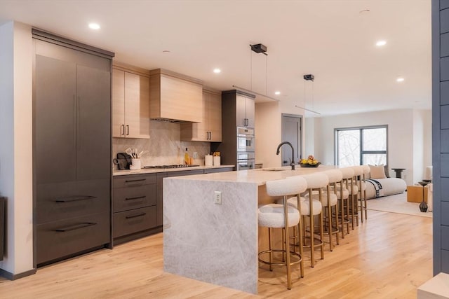 kitchen with pendant lighting, backsplash, premium range hood, a kitchen island with sink, and light wood-type flooring