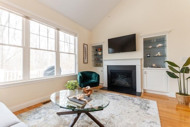 living area featuring wood finished floors, visible vents, baseboards, vaulted ceiling, and a glass covered fireplace