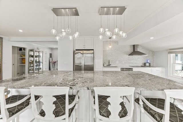 kitchen with stainless steel refrigerator, wall chimney range hood, a sink, tasteful backsplash, and light stone countertops