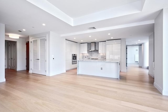 kitchen featuring backsplash, a kitchen island with sink, light hardwood / wood-style flooring, wall chimney exhaust hood, and white cabinetry