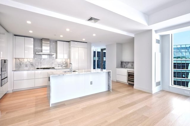 kitchen with white cabinets, wall chimney exhaust hood, an island with sink, and wine cooler