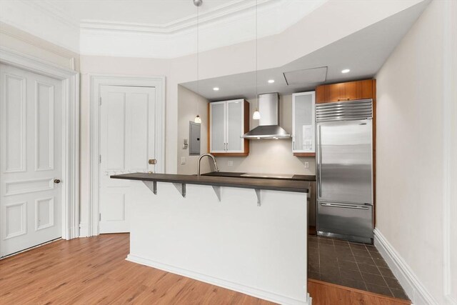 kitchen featuring wall chimney range hood, dark countertops, stainless steel built in refrigerator, and a breakfast bar area