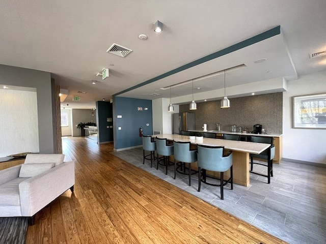 kitchen featuring an island with sink, backsplash, stainless steel refrigerator, a kitchen bar, and decorative light fixtures