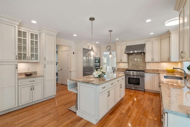 kitchen with wall chimney exhaust hood, high end appliances, a sink, light wood-style flooring, and recessed lighting
