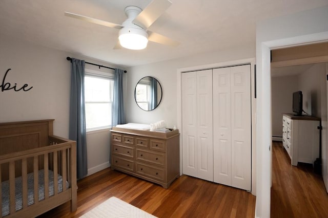 bedroom featuring ceiling fan, a crib, baseboard heating, wood finished floors, and a closet