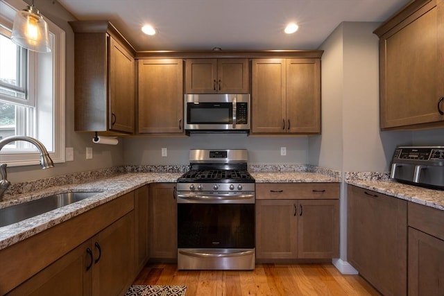 kitchen with sink, hanging light fixtures, stainless steel appliances, light stone counters, and light hardwood / wood-style flooring