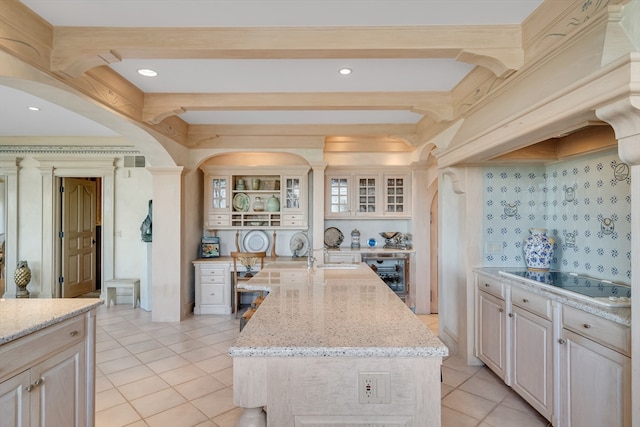 kitchen with beverage cooler, a kitchen island with sink, stovetop, decorative columns, and light stone countertops