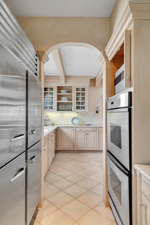 kitchen with beam ceiling, stainless steel double oven, light brown cabinets, and light tile patterned floors