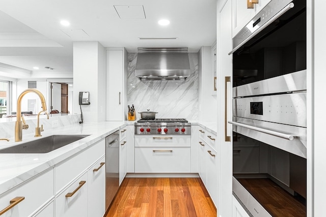 kitchen featuring sink, wall chimney range hood, stainless steel appliances, light stone counters, and white cabinets