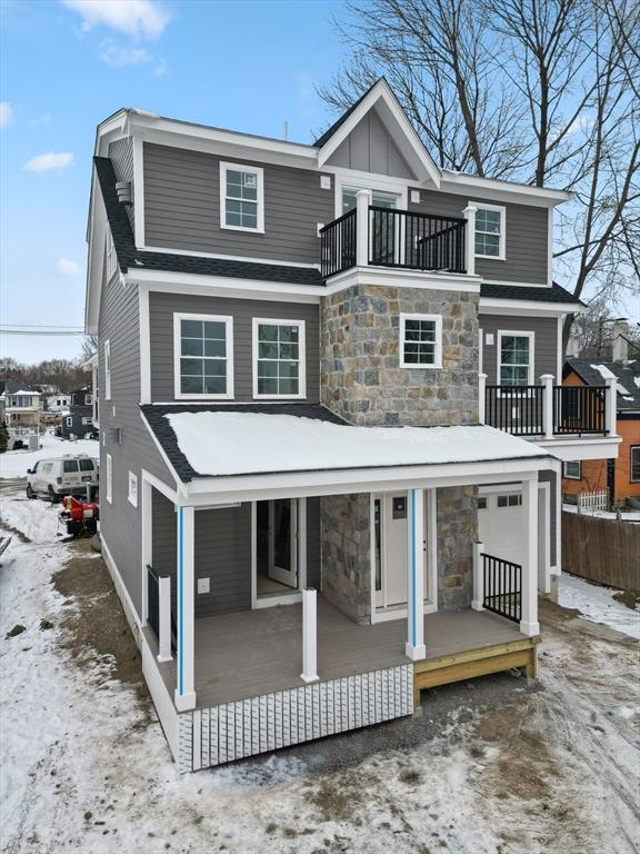 snow covered house with covered porch, board and batten siding, a balcony, and stone siding