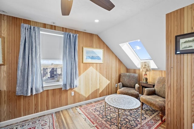 living area with ceiling fan, hardwood / wood-style floors, lofted ceiling with skylight, and wood walls