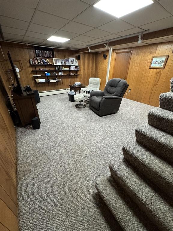 living area with carpet, wooden walls, a paneled ceiling, a baseboard heating unit, and stairs