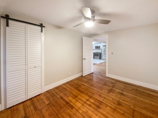 unfurnished bedroom with stainless steel refrigerator, ceiling fan, a barn door, a closet, and hardwood / wood-style flooring