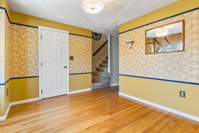 empty room featuring stairway, wood finished floors, baseboards, wallpapered walls, and wainscoting