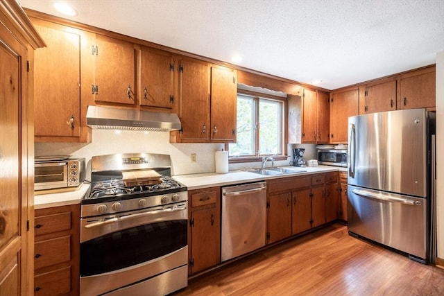 kitchen featuring appliances with stainless steel finishes, wall chimney exhaust hood, a sink, light wood-style floors, and brown cabinetry