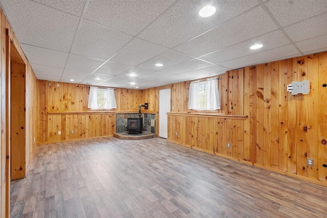 unfurnished living room with wood finished floors, wood walls, baseboards, a wood stove, and a paneled ceiling