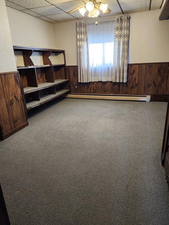 empty room with ceiling fan, a baseboard radiator, carpet floors, a paneled ceiling, and wooden walls