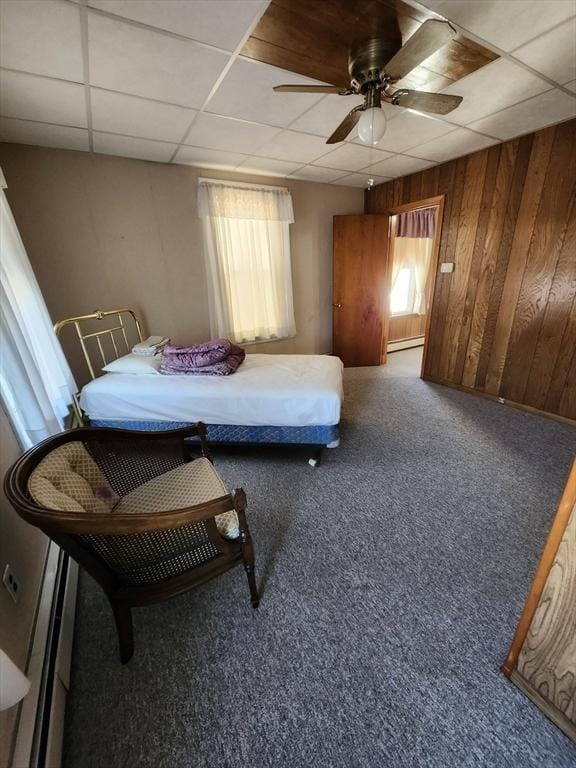 carpeted bedroom with a baseboard heating unit, a paneled ceiling, ceiling fan, and wooden walls