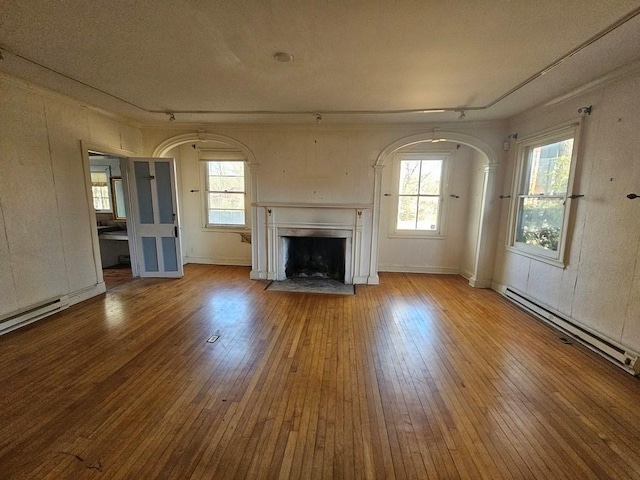 unfurnished living room with a baseboard heating unit, arched walkways, wood-type flooring, and a fireplace with flush hearth