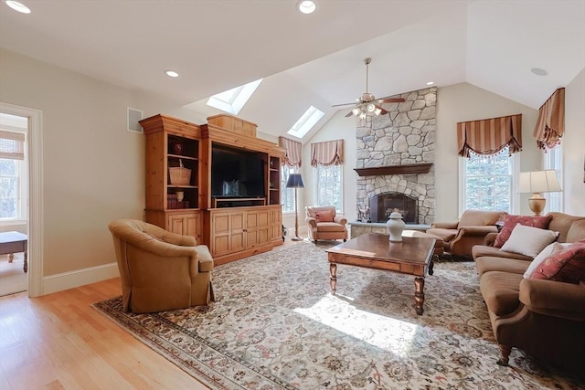 living room featuring light wood-type flooring, lofted ceiling with skylight, baseboards, a ceiling fan, and a stone fireplace