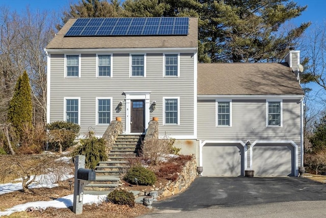 colonial house featuring roof mounted solar panels, driveway, a chimney, and an attached garage