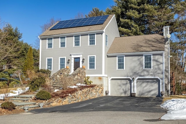 colonial home featuring an attached garage, a shingled roof, driveway, roof mounted solar panels, and a chimney