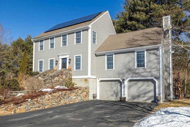 colonial home featuring a garage, roof mounted solar panels, driveway, and a chimney