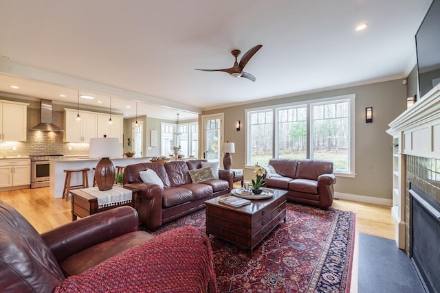 living area with ceiling fan, a tiled fireplace, baseboards, light wood finished floors, and ornamental molding
