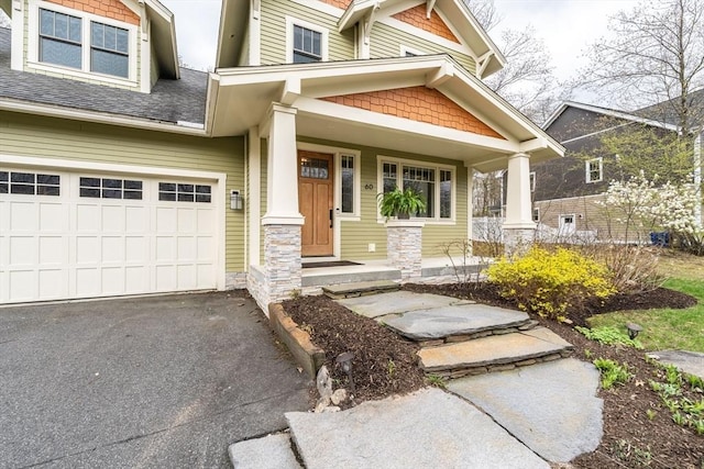 view of front facade with covered porch, driveway, an attached garage, a shingled roof, and stone siding