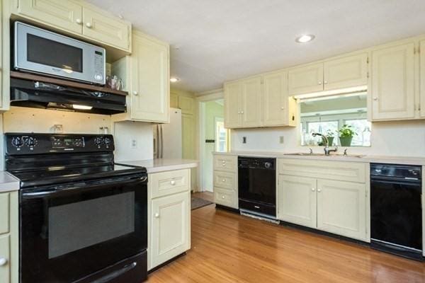 kitchen with light countertops, black appliances, under cabinet range hood, a sink, and wood finished floors