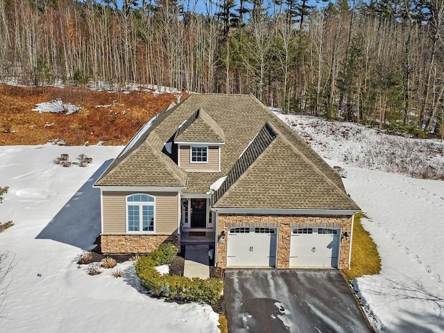 view of front of home featuring an attached garage, a shingled roof, stone siding, driveway, and a view of trees