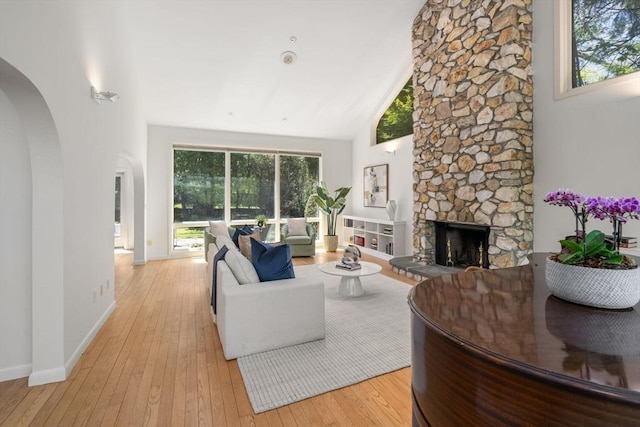 living room with a stone fireplace, arched walkways, baseboards, high vaulted ceiling, and light wood-style flooring