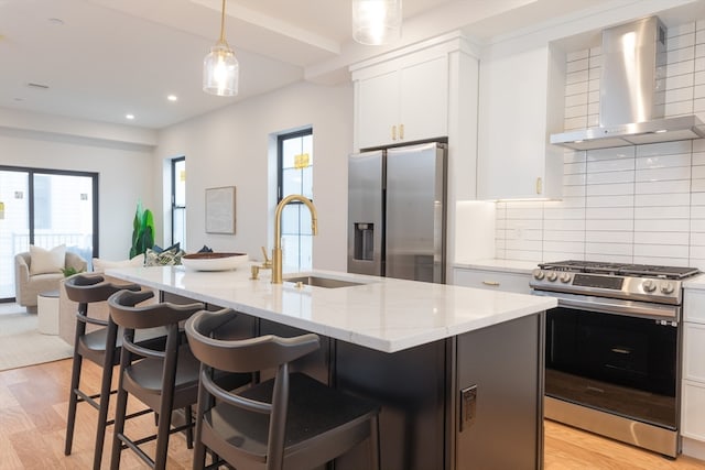 kitchen with an island with sink, white cabinets, stainless steel appliances, and wall chimney range hood